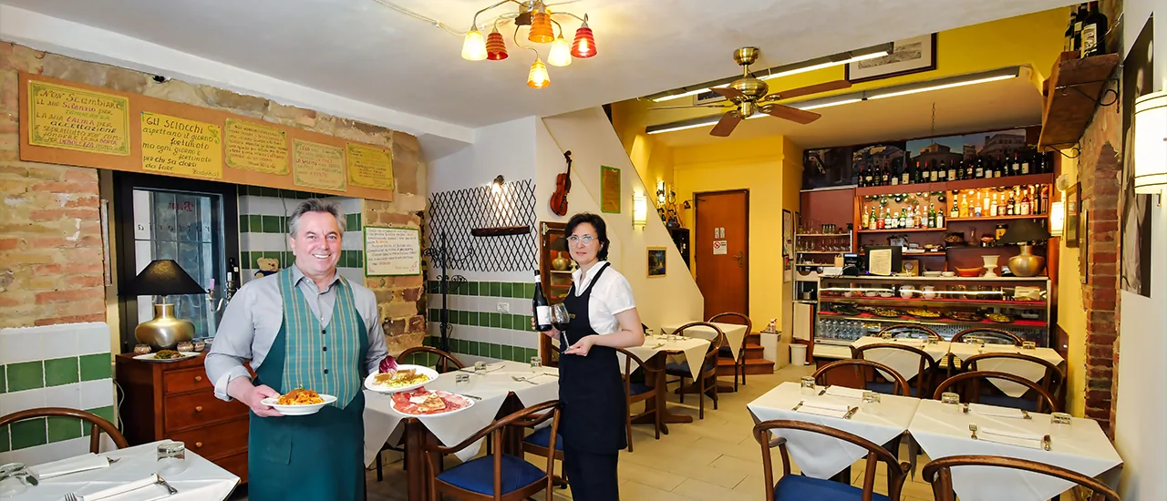 Lolita e Vittorio nella sala di Bottega Dei Priori, con piatti preparati in cucina e una bottiglia di vino.
